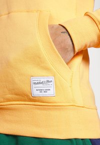 Żółta bluza z kapturem z prążkowanymi mankietami i dołem, z kieszenią z przodu oraz metką z napisem "Mitchell & Ness" dla identyfikacji marki.