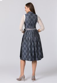 Blau gemustertes Kleid mit tailliertem Oberteil und gefälteltem Rock. Besitzt Spitzenärmel und ein gebundenes Taillendetail. Graue High-Heels.