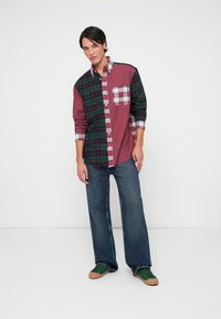 Camicia a scacchi con pannelli rossi e verdi, con una tasca quadrettata. Indossata con jeans a gamba larga blu e sneakers verdi.