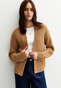 Brun stickad cardigan med ett strukturerat mönster, öppen front och långa ärmar. Bärs över en vit topp och matchas med mörka jeans.
