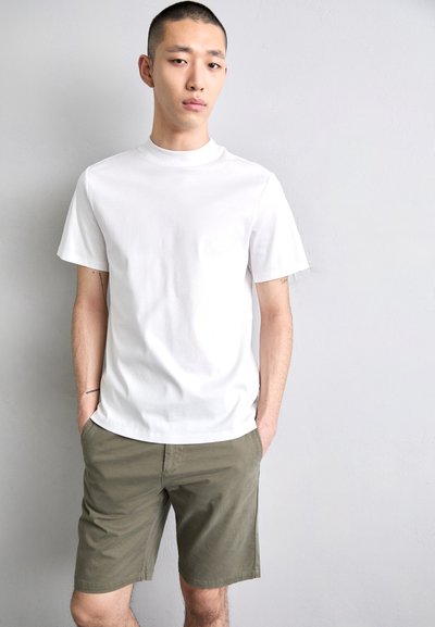 T-shirt en coton blanc à manches courtes et col rond, associé à un short vert olive. Le mannequin adopte une posture neutre avec les mains dans les poches.