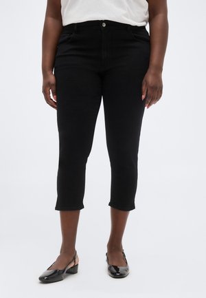 Person iført sorte korte bukser og sorte lavhælede slingback-sko, stående mod en ensartet lys baggrund.