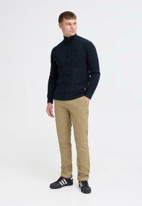 Pull de couleur bleu marine foncé avec un col montant et une fermeture éclair quart, associé à un pantalon chino couleur kaki et des baskets noires avec des rayures blanches.