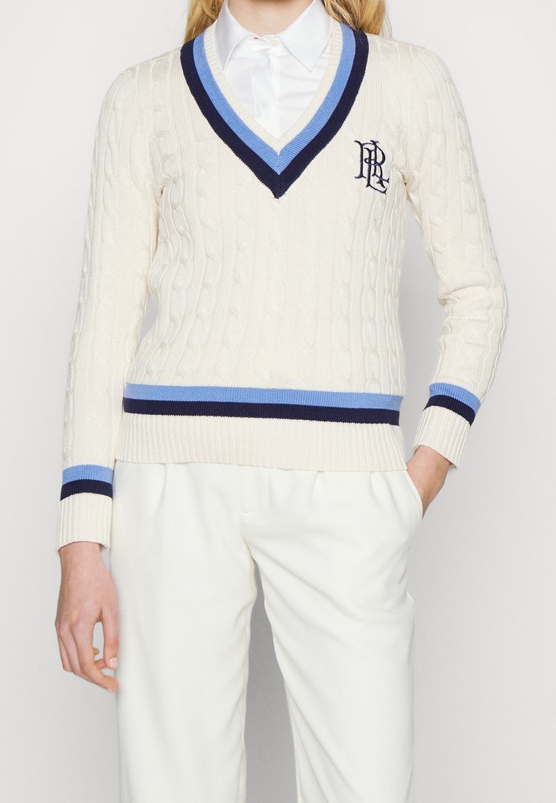 Femme portant un pull en maille torsadée crème avec un col en V rayé bleu marine et bleu, poignets et bas assortis, associé à un pantalon blanc et une chemise à col.