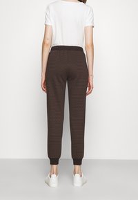 Kvinna i mörkbruna mönstrade joggers, vit t-shirt och vita sneakers, står mot en enkel ljus bakgrund vänd bort.