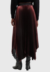Gonna plissé rossa con orlo alto-basso e vita nera, caratterizzata da pieghe verticali e una texture liscia e brillante, indossata sopra stivali alti fino al ginocchio.