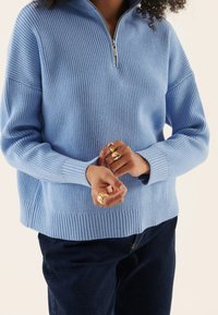 Eine Person, die einen hellblauen Reißverschluss-Pullover trägt, zeigt mehrere goldene Ringe an ihren Händen, vor einem neutralen Hintergrund.