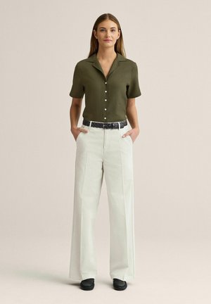 Kvinde stående med hænderne i lommerne iført en olivengrøn kortærmet skjorte med knapper, hvide vidde bukser, sort bælte og sorte sko.