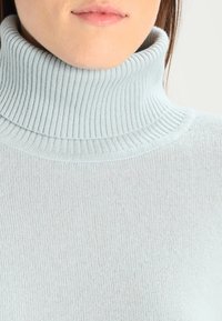 Frau in einem hellgrauen, gerippten Rollkragensweater, zeigt Hals und Lippen vor einem schlichten Hintergrund.