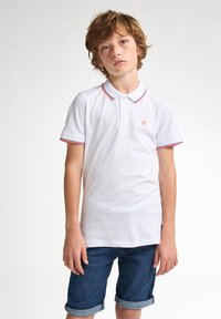 Valkoinen polo paita lyhyillä hihoilla, punaiset korostusraidat kauluksessa ja hihoissa sekä pieni logo rinnassa. Yhdistetty denim-shortsien kanssa.