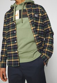 Camicia di flanella con motivo a quadri verde, giallo e blu navy; presenta una tasca sul petto e chiusura con bottoni sopra un hoodie verde con un logo.