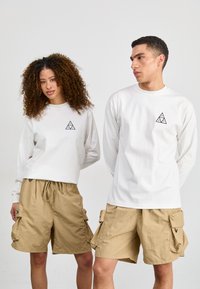 Hvite langermede skjorter med en triangulær svart logo, parret med beige cargo shorts med sidelommer og snøring i livet.