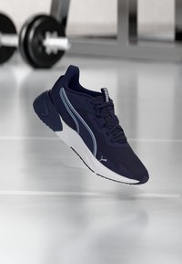 Σκούρο μπλε αθλητικό παπούτσι με λευκή σόλα και ρίγες στο πλάι, αιωρούμενο πάνω από καθρεφτισμένο δάπεδο γυμναστηρίου με θολό αλτήρα στο φόντο.