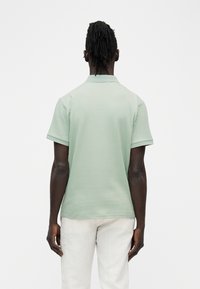 Homme à la peau foncée et aux dreadlocks portant un polo à manches courtes côtelé vert clair et un pantalon blanc, debout de dos sur un fond blanc.