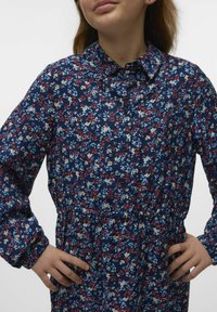 Abito floreale blu navy con colletto a bottoni, vita elastica e maniche lunghe che presentano motivi floreali bianchi, rossi e azzurri.