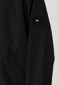Schwarze Windbreaker-Jacke mit feiner Textur, Knopfverschluss und einem Patch mit dem "TOMMY HILFIGER"-Logo sowie farblichen Akzenten am Arm.