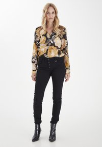 Pantalones negros con cierre de botón frontal y costuras laterales, combinados con una blusa floral que presenta rosas amarillas y blancas sobre un fondo negro.