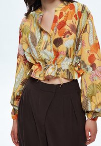 Blusa tagliata, botton-up in giallo con motivi floreali in rosso, arancione e verde. Presenta maniche lunghe con polsini arricciati e un dettaglio a fiocco all'orlo.
