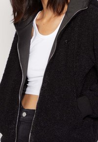 Czarna bluza z polaru zapinana na zamek, z teksturowaną powierzchnią zewnętrzną, szarą podszewką, ściągaczami na rękawach i kieszeniami. Noszona na białym krótkim topie.