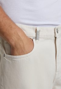 Beige cotton pants with a smooth texture, featuring a side pocket and a metal tag at the waistband. Hand positioned in the pocket.