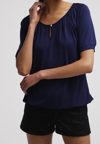 Mörkblå kortärmad topp med rundad halsringning och en nyckelhålsdetalj, tillsammans med texturerade svarta shorts.