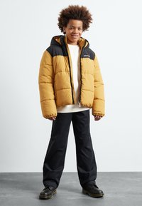 Veste matelassée jaune avec une section supérieure noire, fermeture éclair à l'avant et capuche, portée avec un pantalon noir et des chaussures noires.