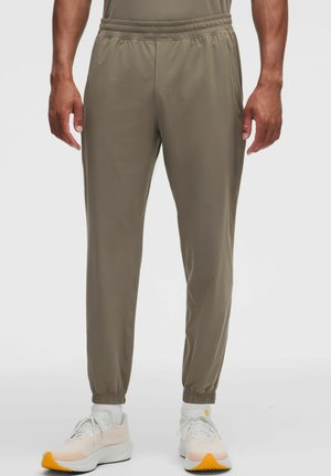 Man staand, draagt olijfgroene joggerbroeken met een elastische taille en boorden, gecombineerd met witte sportschoenen met oranje zolen.