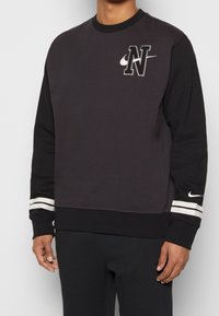 Man som bär en svart och mörkgrå Nike-tröja med lång ärm, vita ränder på ärmarna och en sydd "N"-logotyp på bröstet.