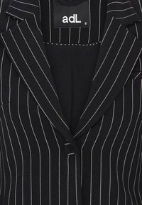 Mustat raitapuvutakki, joissa on valkoiset pystyrakenteet, kaulukset, yksittäinen napitus ja näkyvä adL-brändilogo kauluksessa.