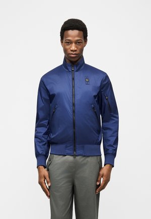 Jeune homme debout, portant une veste bomber bleu marine zippée avec des poches zippées et un pantalon gris, sur un fond uni.