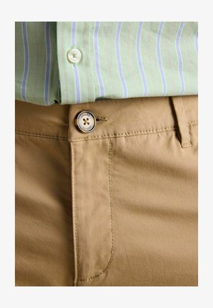 Khakihose mit einem Knopf mit der Aufschrift "Joules", kombiniert mit einem eingesteckten grün gestreiften Hemd mit weißen und blauen Linien.