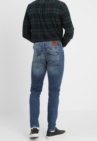 Blå denimjeans med slim fit, prydda med slitna detaljer och bakfickor, i kombination med en mönstrad flanellskjorta och svarta sneakers.