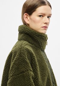 Õlivihreast fliisist jakk kõrge kraega, tekstuurse pinnaga, tõmblukuga ja sileda voodriga sees. Zipperil on rõngastõmmits.