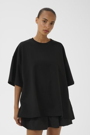 Donna con capelli tirati indietro che indossa una camicia nera oversize a maniche corte e pantaloncini neri su uno sfondo chiaro uniforme.