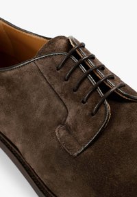 Zapato de ante marrón con una textura suave, que presenta un diseño de cordones cerrado y costuras negras contrastantes a lo largo del borde y los cordones.