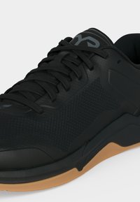 Μαύρο αθλητικό παπούτσι με διχτυωτό επάνω μέρος, μαύρα κορδόνια και σόλα σε χρώμα καουτσούκ, με το λογότυπο της μάρκας στη γλώσσα.