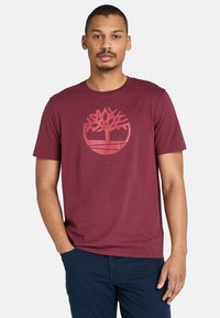 Burgundy t-shirt i bomull med en cirkulär trälogga i en ljusare nyans. Kort ärm, rund halsringning och en avslappnad passform.