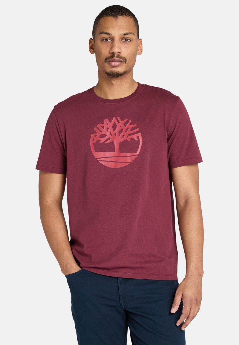 Burgundy t-shirt i bomull med en cirkulär trälogga i en ljusare nyans. Kort ärm, rund halsringning och en avslappnad passform.