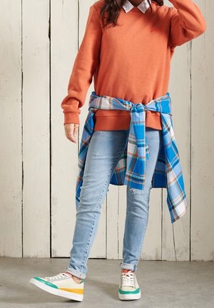 Suéter naranja con cuello, camisa de cuadros azul claro atada a la cintura, jeans azules desgastados y zapatillas multicolores.