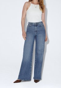 Femme portant un haut blanc sans manches, un jean bleu taille haute à jambes larges, et des chaussures marron pointues, debout devant un fond uni.