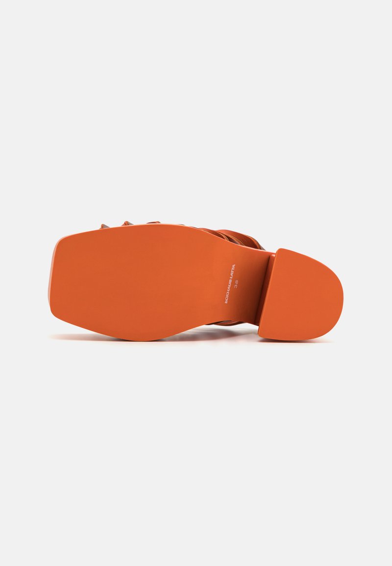 Sinaasappel kleurige hak met een gladde, solide zool en een ontwerper met bandjes bovenaan. Het materiaal lijkt synthetisch met een matte afwerking.