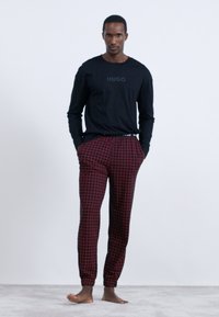 Schwarzes Langarmshirt mit "HUGO"-Logo, kombiniert mit rot-karierten Schlafanzughosen mit elastischen Bündchen. Glatte Texturen durchgehend.