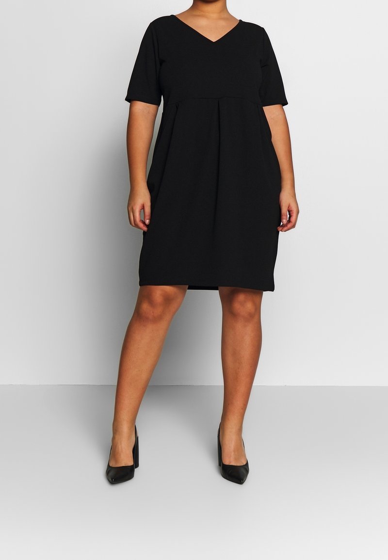 Schwarzes Kleid mit V-Ausschnitt, kurzen Ärmeln und gerafftem Taillendetail. Geschmeidiger Stoff und knielanger Schnitt. Getragen mit schwarzen High-Heels.