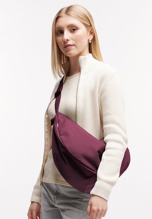 Giovane donna con capelli biondi che indossa un maglione crema con cerniera, pantaloni grigi e porta una grande borsa a tracolla viola.