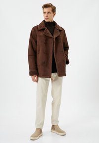 Chaqueta marrón de doble botonadura con cuello y puños de borreguillo, hecha de material suave. Combinada con pantalones beiges y zapatos de tela de color claro.