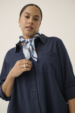 Žena u mornarsko plavoj košulji s podvrnutim rukavima, prugastim plavo-bijelim šalcem oko vrata, srebrnim naušnicama i prstenjem na prstima.