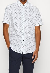 Camicia bianca a maniche corte realizzata in tessuto con un motivo blu, colletto button-down, bottoni blu navy e un piccolo logo sul petto.