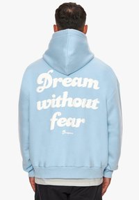 Hellblaue Kapuzenjacke aus weichem Material. Mit auffälligem weißen Text "Träume ohne Angst" auf dem Rücken und einer Kängurutasche vorne.