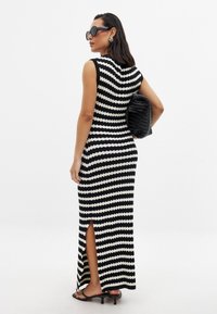 Robe maxi en tricot rayée en noir et blanc avec une coupe ajustée, fente sur le côté et sans manches. Accessoire : pochette noire texturée.