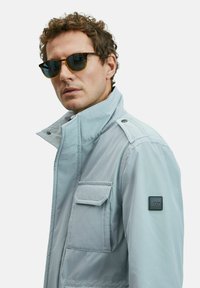 Man wearing light gray jacket with flap pocket and sunglasses, looking slightly to the side against a plain background.